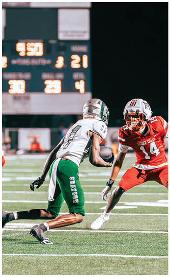 A person in a white and green football uniform is running with the ball on a field while two people in red uniforms approach from either side. Description generated by AI