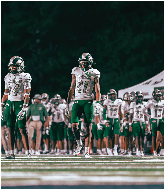 A group of people in football uniforms with helmets are standing on a field, with one person prominently in the foreground wearing a jersey numbered 21. Description generated by AI