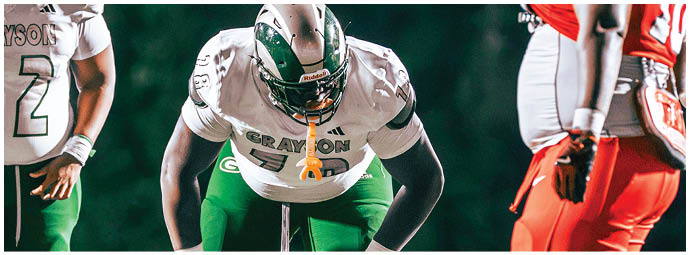 Three people in football uniforms are positioned on a field, with one person in a white jersey and green pants bent forward in a ready stance between two others wearing green and red pants. Description generated by AI
