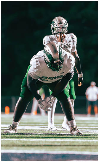 Two people in football uniforms and helmets are positioned on a field with one crouching and holding a football while the other stands behind. Description generated by AI