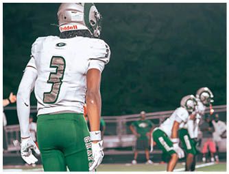 A person wearing green sports shorts with the word “GRAYSON" on the side and white gloves stands on a football field with other players in the background. Description generated by AI