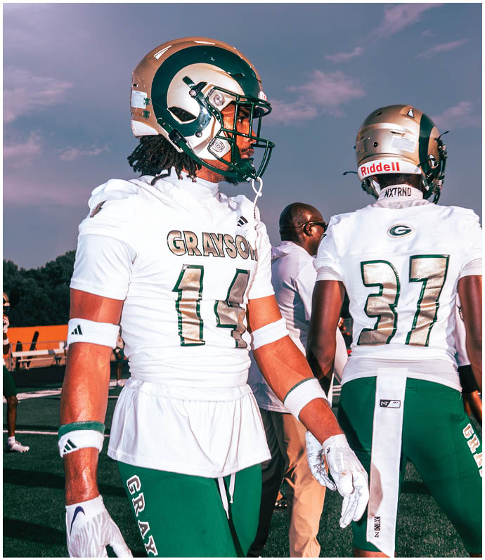 Two people in football uniforms with helmets stand on a field, one facing forward and the other facing away, with a third person in casual clothing in the background. Description generated by AI