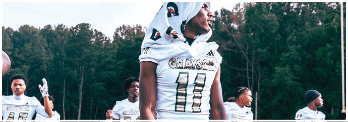 People wearing football uniforms are standing outdoors on a field with trees in the background, with one person in the center wearing a jersey with the name “GRAYSON" and the number 11. Description generated by AI