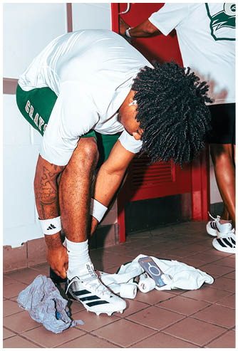 A person wearing a white shirt, green shorts, and white Adidas socks is bending down to tie their white Adidas sneakers while wearing wireless earbuds. Description generated by AI