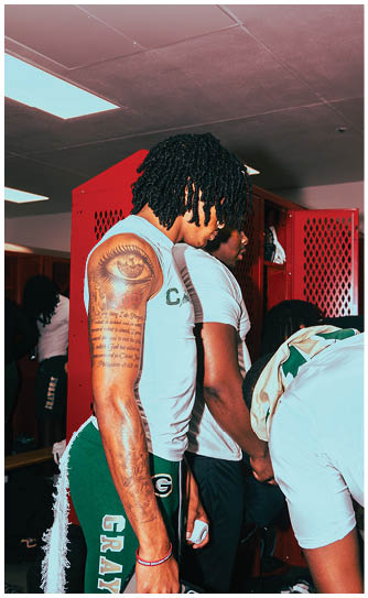 A person with curly hair wearing a light blue sleeveless top stands near a red locker in an indoor setting with fluorescent ceiling lights. Description generated by AI