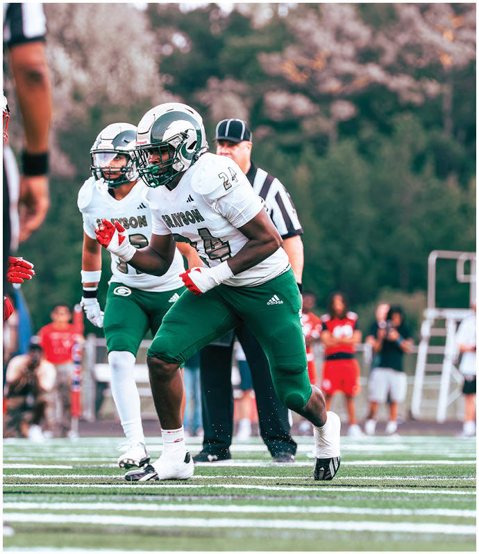 Two people in football uniforms and helmets are on a field with a referee standing behind them. Description generated by AI
