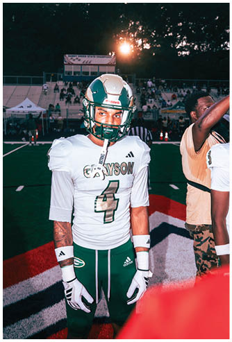A person wearing a football helmet is in the foreground with a stadium and a scoreboard reading “Sandy Creek State Champions" in the background during sunset. Description generated by AI
