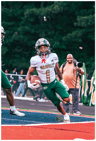 A person in a white and green football uniform with the name Grayson is running on a field holding a football while another person in similar attire stands nearby and a photographer captures the moment in the background. Description generated by AI