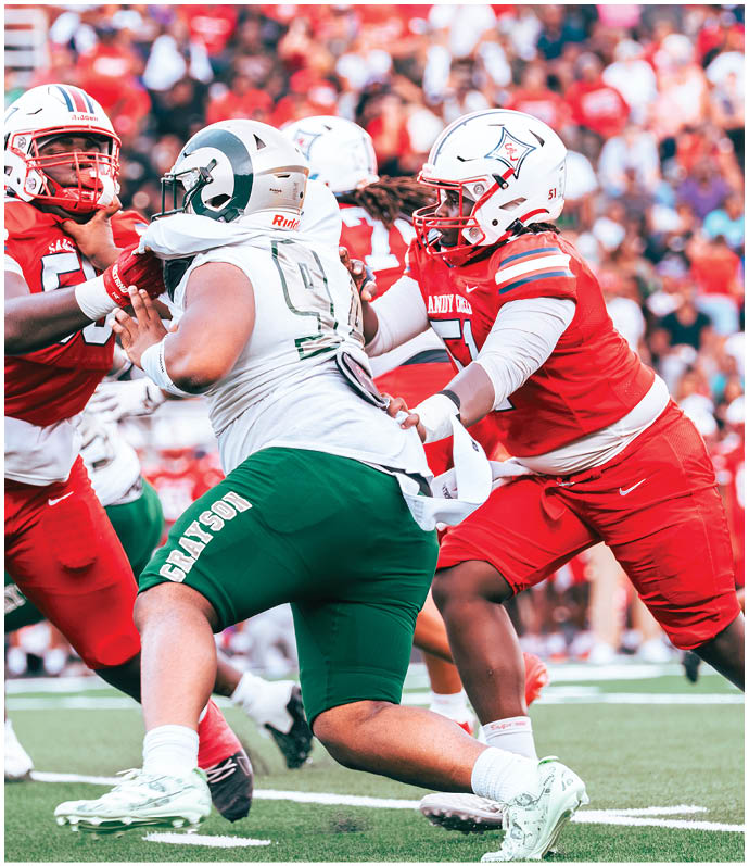 People in red and white football uniforms are actively blocking and tackling a player in a white and green uniform on a crowded field. Description generated by AI