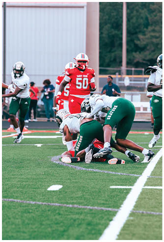 Players in red and white uniforms stand on a football field while players in green and white uniforms tackle an opponent on the ground during a game. Description generated by AI