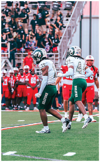 Two people in white and green football uniforms are on a field with a crowd of people in red uniforms and spectators in the background. Description generated by AI