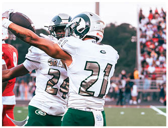 Two people in white and green football uniforms celebrate on a field with one holding a football and a crowd in the background. Description generated by AI