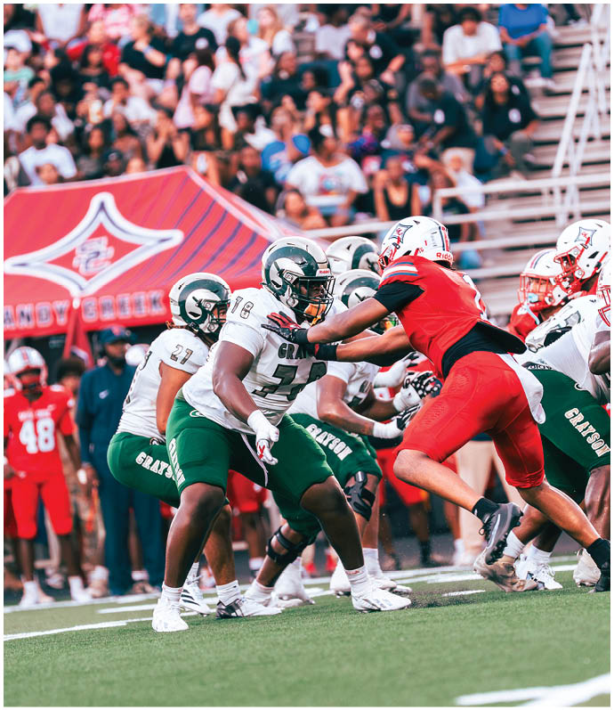 Players in green and white uniforms are blocking a player in a red uniform during a football game with a crowd watching in the background. Description generated by AI