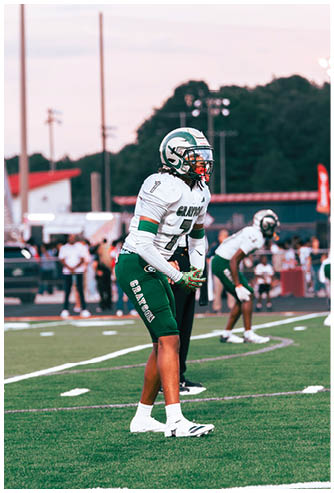 A person in a football uniform with a helmet and gloves is standing on a football field in a ready position during a game. Description generated by AI