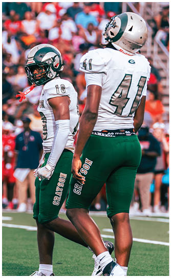 Two people in football uniforms with helmets stand on a field during a game, with one person facing forward and the other partially visible from behind. Description generated by AI
