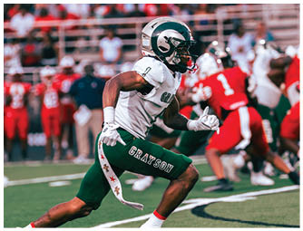 A person wearing a white and green football uniform with the word Grayson on the pants is running on a football field during a game with other players and a crowd in the background. Description generated by AI