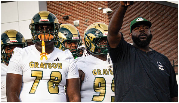 A group of people wearing football helmets and uniforms stand next to a person in a dark shirt and cap who is pointing upwards while speaking. Description generated by AI