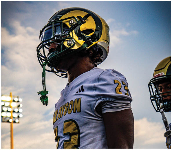 A person wearing a white sports jersey with the number 23 and a gold and black football helmet is looking upward against a cloudy sky. Description generated by AI