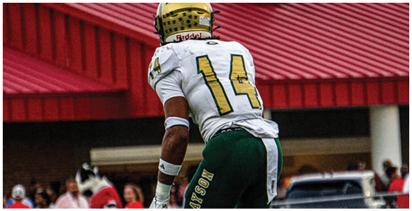 A person wearing a gold football helmet and white jersey with yellow numbers is standing in front of a red metal roof and a building. Description generated by AI