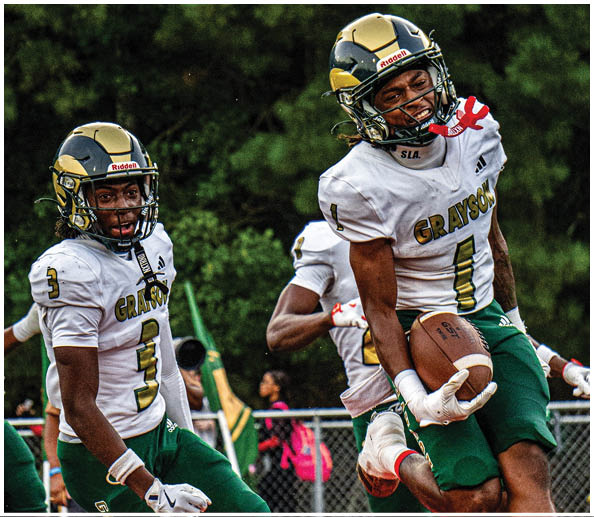 Two people wearing football helmets and jerseys are engaged in a game with a background of green trees. Description generated by AI