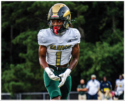 A person wearing a white football jersey with “GRAYSON" and the number one on it, a gold and black helmet, and white gloves is standing outdoors with a green, blurred background. Description generated by AI