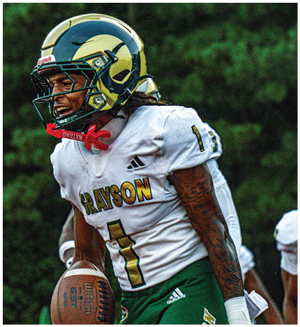 A person wearing a blue and yellow football helmet with a green faceguard is shown against a blurred green background. Description generated by AI