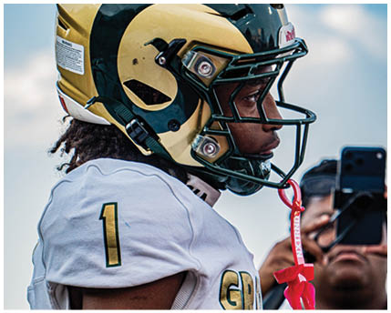 A person wearing a gold and dark green football helmet with a ram horn design is shown in profile against a blue sky background. Description generated by AI