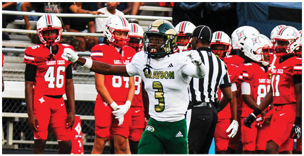 A group of people in football uniforms are on a field with one person in a white jersey gesturing while others in red jerseys and helmets stand nearby and spectators sit in bleachers behind them. Description generated by AI