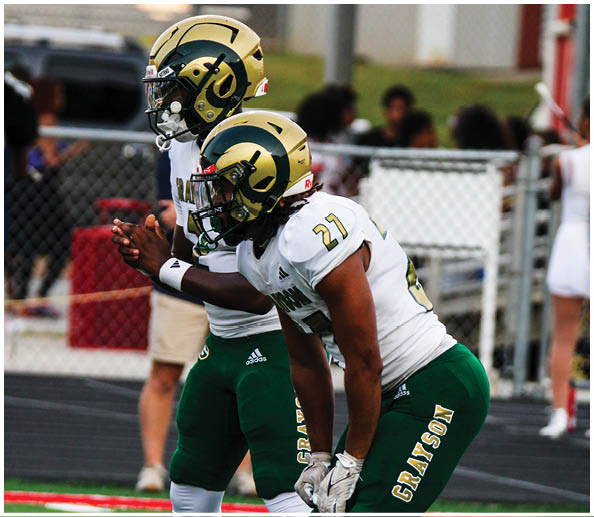 Two people in football uniforms and helmets are positioned as if ready to start a play on a field with a chain-link fence and blurred spectators in the background. Description generated by AI