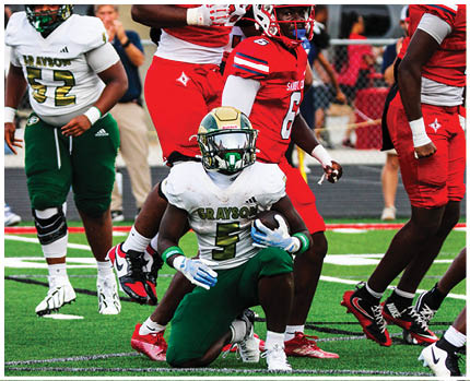 A person in a white and green football uniform is holding a football while crouching on a field with other players in red and white uniforms standing nearby. Description generated by AI