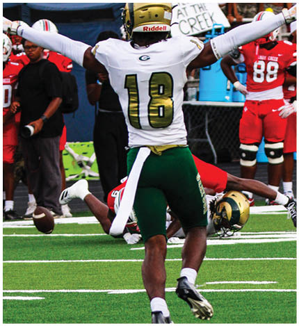A football player wearing a white jersey with the number 18 and green pants raises their arms while another player in red lies on the ground near a loose football on a field. Description generated by AI