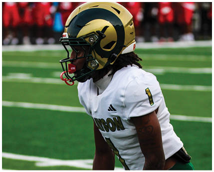 A person wearing a white football jersey with the number one and a gold helmet with a dark green ram horn design is on a football field with blurred players in red uniforms in the background. Description generated by AI