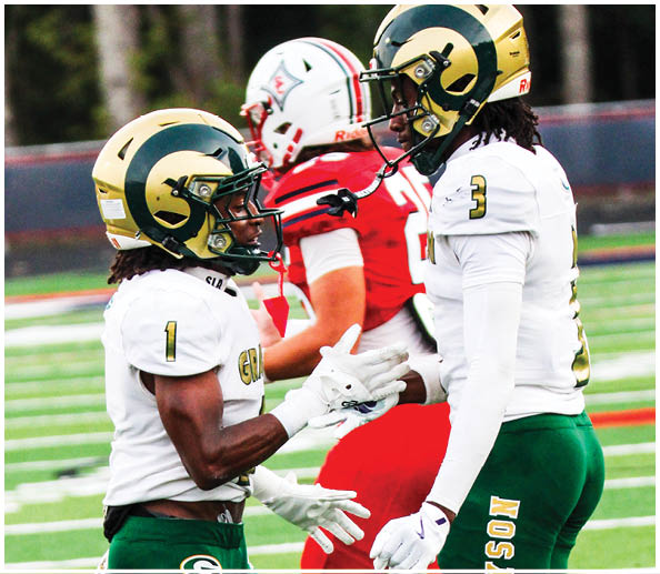 Two people wearing white football uniforms with green and gold helmets are interacting on a football field while a person in a red uniform is in the background. Description generated by AI