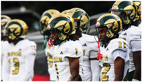 A group of people wearing white football uniforms and gold helmets stand closely together on a field. Description generated by AI