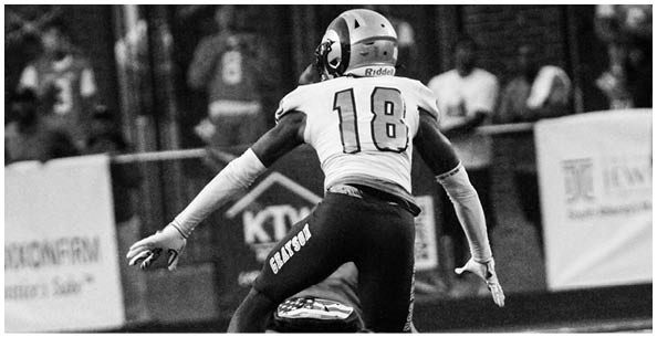 A person wearing a football uniform with the number 18 is in an athletic stance on a field with blurred spectators in the background. Description generated by AI