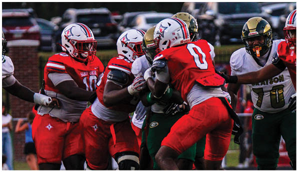 Several people wearing football uniforms are engaged in a close physical play on a field with vehicles visible in the background. Description generated by AI