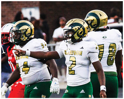 Three people wearing football helmets and uniforms are on a field, with one person pointing while others look in the same direction. Description generated by AI