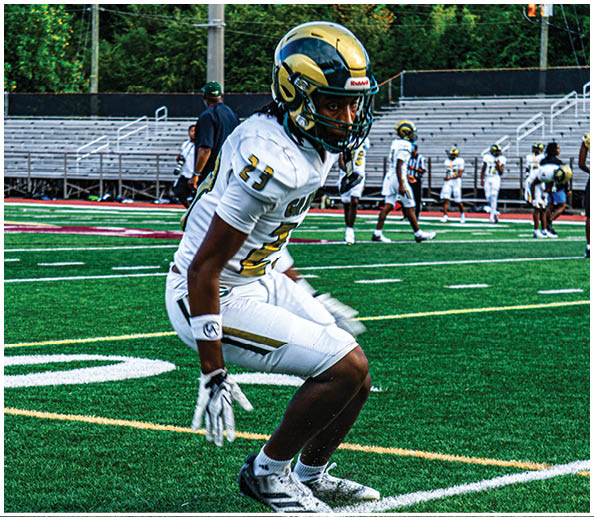 A person in a white and gold football uniform is crouched in an athletic stance on a green football field with other players and a coach in the background. Description generated by AI