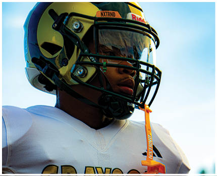 A person wearing a gold and black football helmet and a white jersey with the word “BRAVES" visible is looking to the side against a blue sky background. Description generated by AI