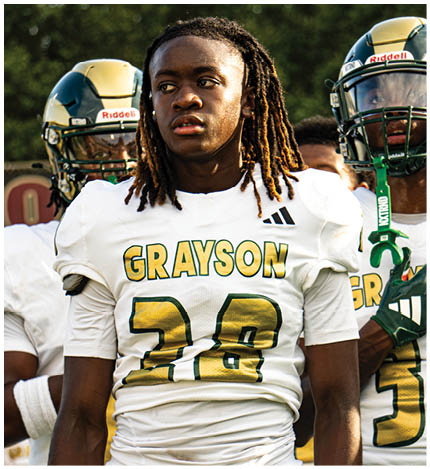 A person with long dreadlocks wearing a white football jersey with the name Grayson and the number 28 stands in front of two other people in similar football uniforms and helmets. Description generated by AI
