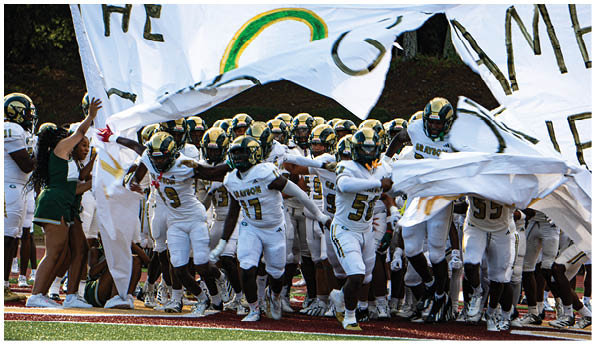 A group of football players in white uniforms and helmets run energetically through a torn banner held by cheerleaders on a field. Description generated by AI