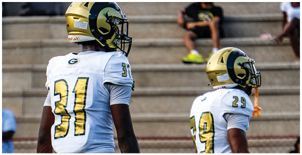 Two people wearing white sports uniforms with green and yellow stripes stand on a field with others in the background. Description generated by AI