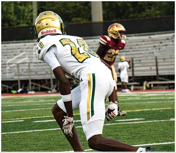 A person in a white and green football uniform with a gold helmet is on a field facing away from the camera while another person in a maroon and gold uniform stands in the background near empty bleachers. Description generated by AI