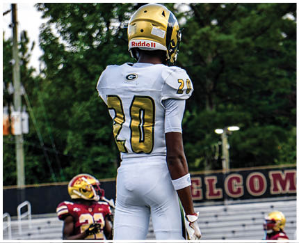 A person wearing a white and gold football uniform with the number 20 stands on a field while two other people in red and gold football uniforms are visible in the background near empty bleachers. Description generated by AI