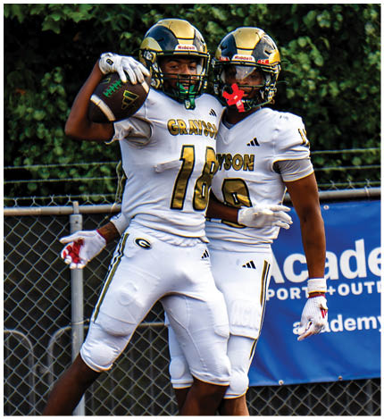 Two people in football uniforms celebrating on a field with a chain-link fence and a blue Academy Sports + Outdoors banner in the background. Description generated by AI
