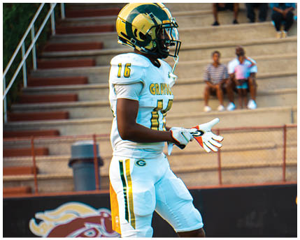 A person wearing a white and yellow football uniform with the number 16 is standing on a field near bleachers where two people are seated. Description generated by AI