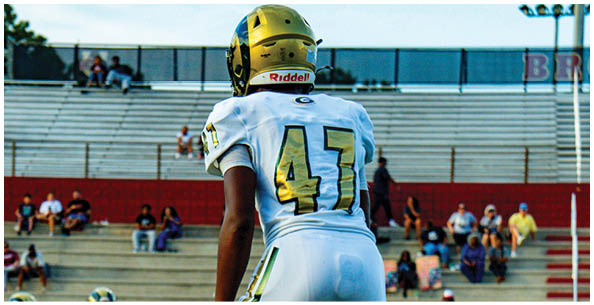 A person wearing a white sports uniform with the number 47 is standing on a field with other people in similar uniforms and spectators seated in bleachers in the background. Description generated by AI