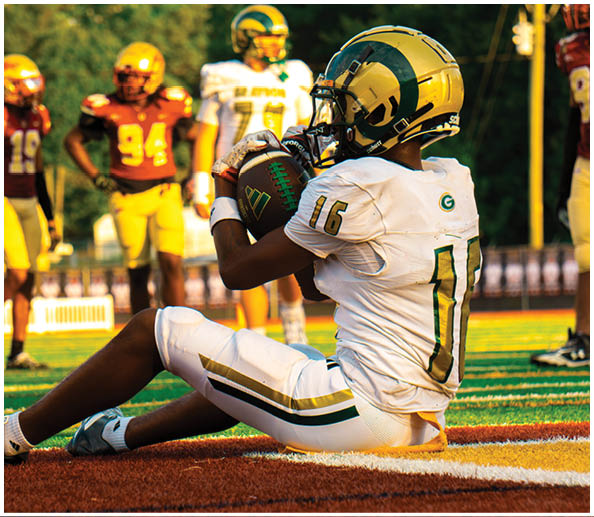 A person in a white and gold football uniform is sitting on the field holding a football while other players stand in the background. Description generated by AI