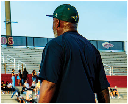 A person wearing a dark shirt and cap stands in front of bleachers with scattered people and a group of cheerleaders performing on a sports field. Description generated by AI