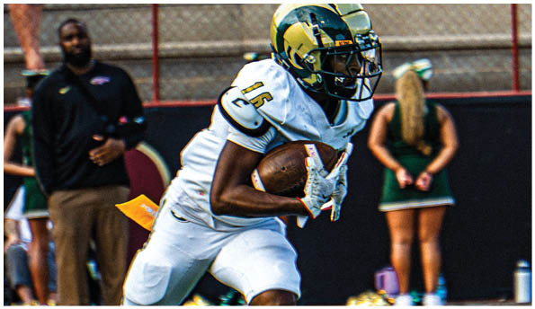 A person wearing a white football uniform and helmet is running with a football on a field while people in green cheerleading outfits stand in the background. Description generated by AI